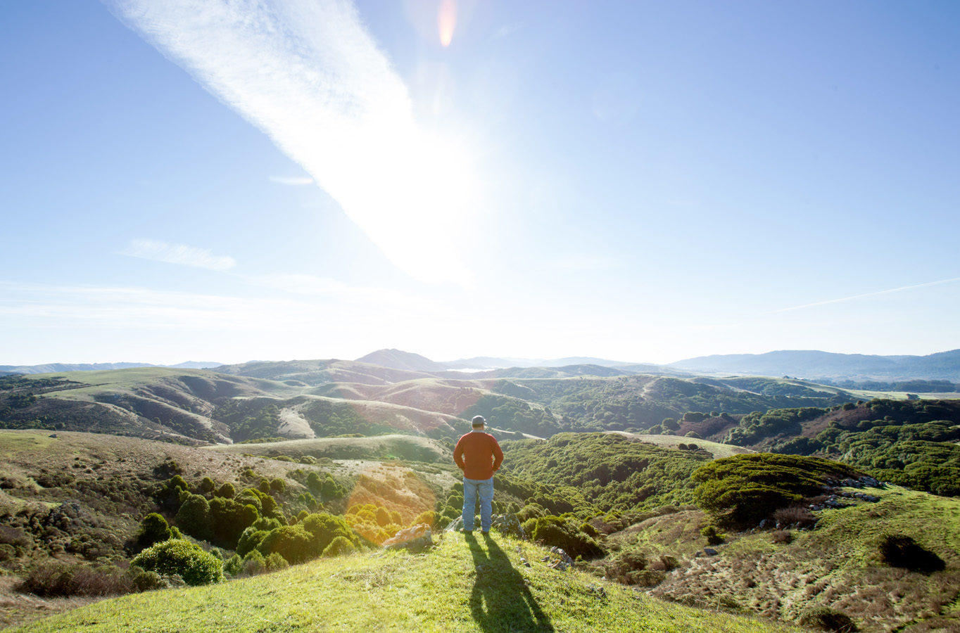 Man atop a green hill, looking out at more rolling hills, sunshine, and blue skies. Photo by Paige Green.