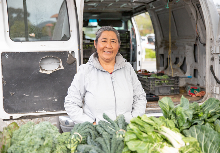 A woman vendor at a farmers' market stands in front of an open van with an array of vegetables on a table in front