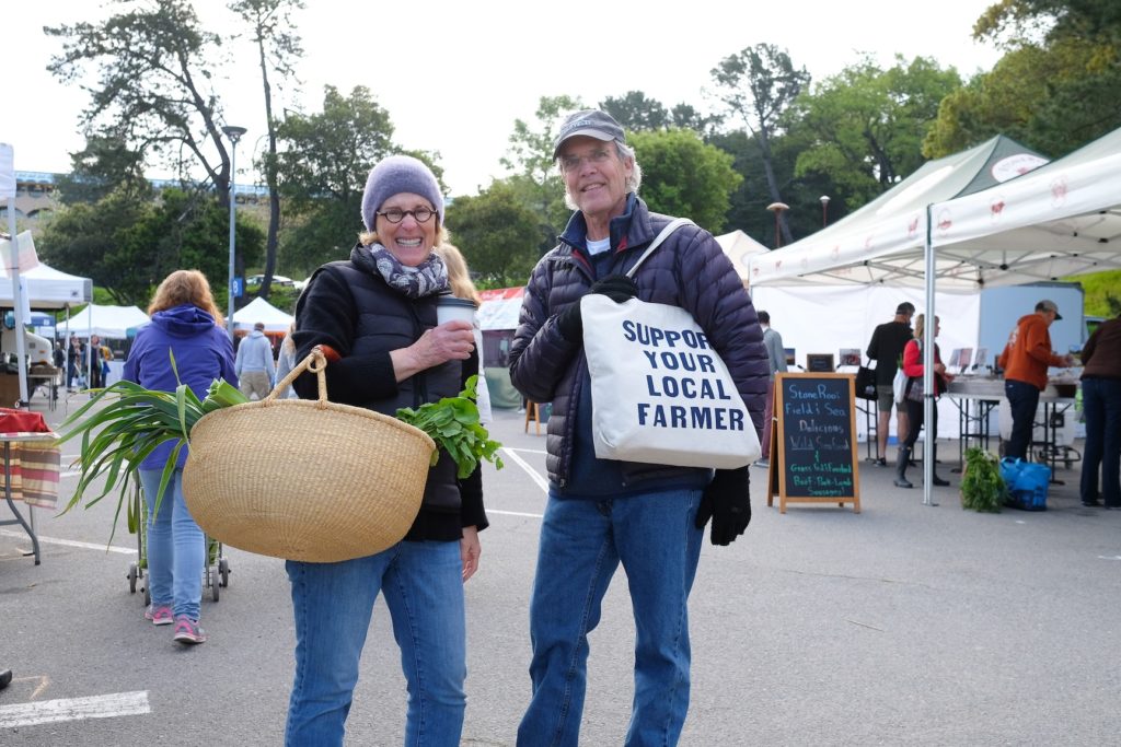 A man and woman carrying a basket and a tote bag that says "Support Your Local Farmer" outside at a farmers' market event. 