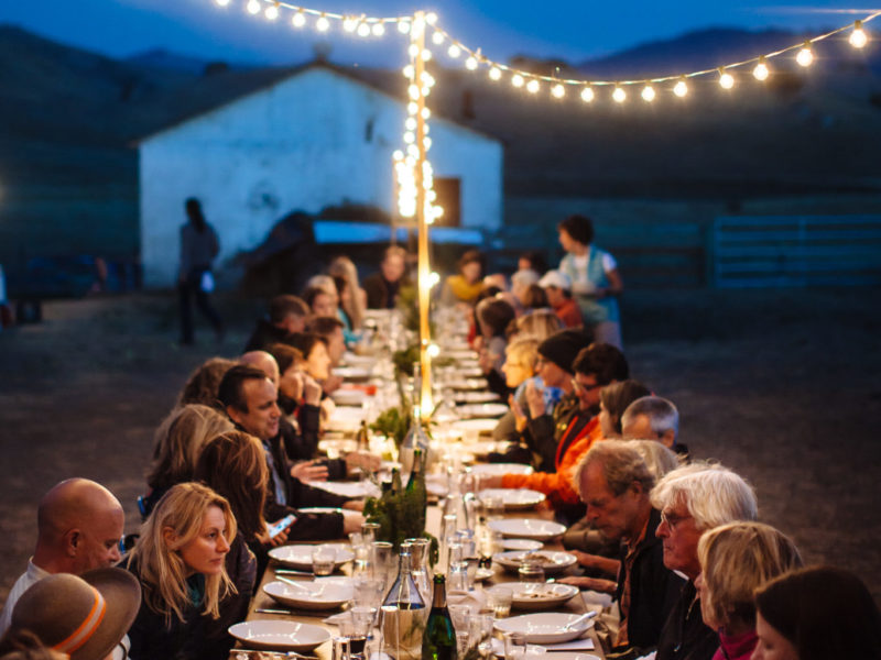 A long dining table full of people sharing food together sits outside in a field in the evening, with a barn and string lights in the background at a field-to-fork event on the land.