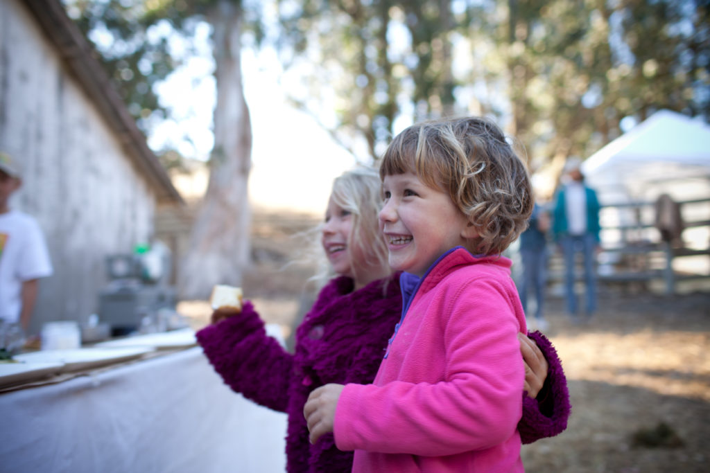 Two young girls in winter coats, watching something out of view, laughing at a family fun event.