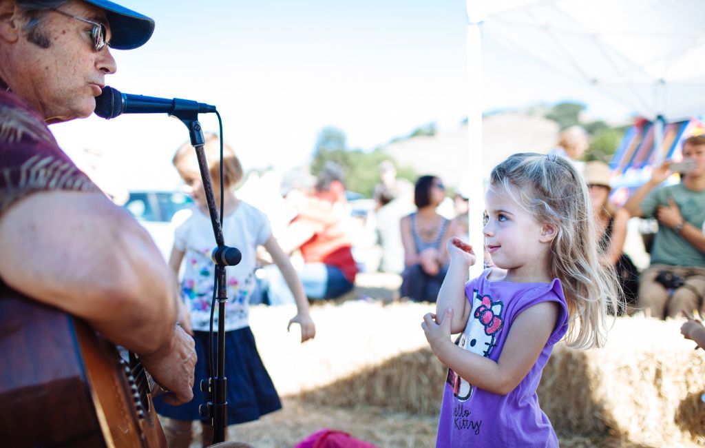 A little girl in a purple shirt stands in front of a seated man playing guitar and singing into a microphone at a family event in Marin. 