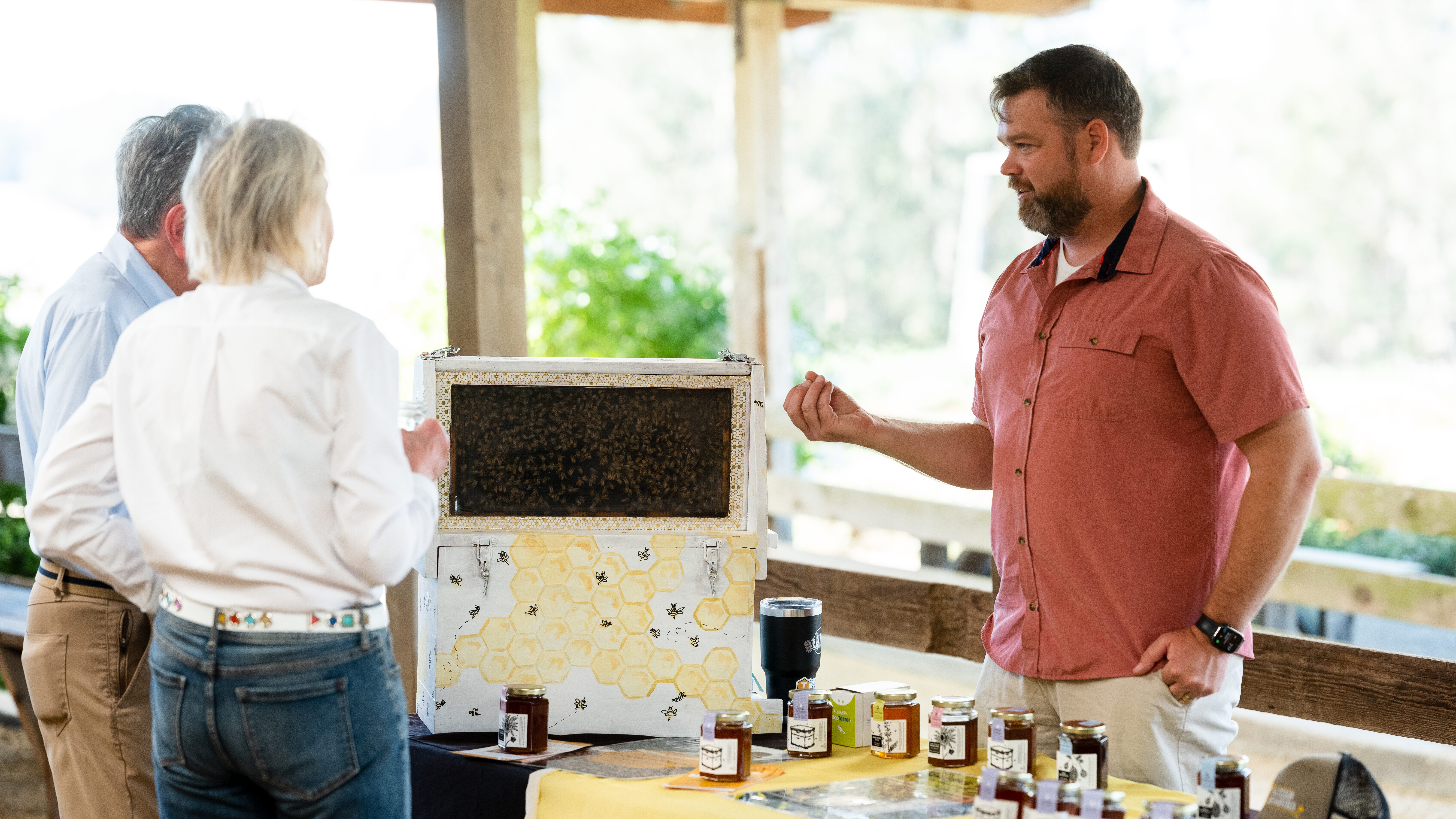 A representative from Sola Bee Honey stands at a booth with jars of honey and a beehive box, talking to two attendees. Photo by Paige Green Photography for MALT.