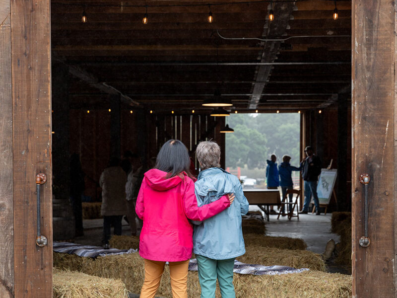 Two women stand at the entrance to a barn with their arms around each other's shoulders.