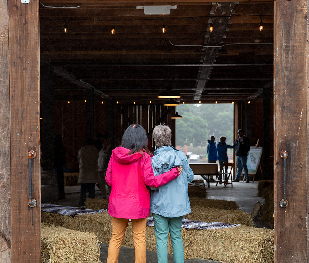 Two women stand at the entrance to a barn with their arms around each other's shoulders.