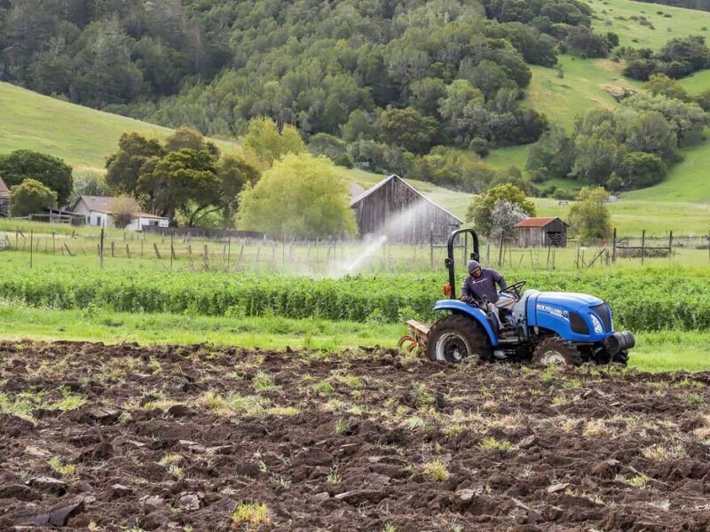 A farmer on a blue tractor plowing a vegetable field against a backdrop of green hills