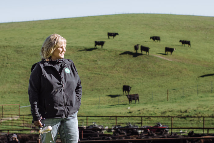 Dayna Ghirardelli stands on green hillside holding a ranch tool, with black cattle grazing on slopes behind her and corrals visible in the middle distance