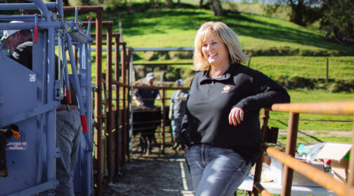 Dayna Ghirardelli leans against wooden corral rails at Duncan Ranch, wearing a black pullover and jeans, with cattle handling equipment and green pastures visible behind her