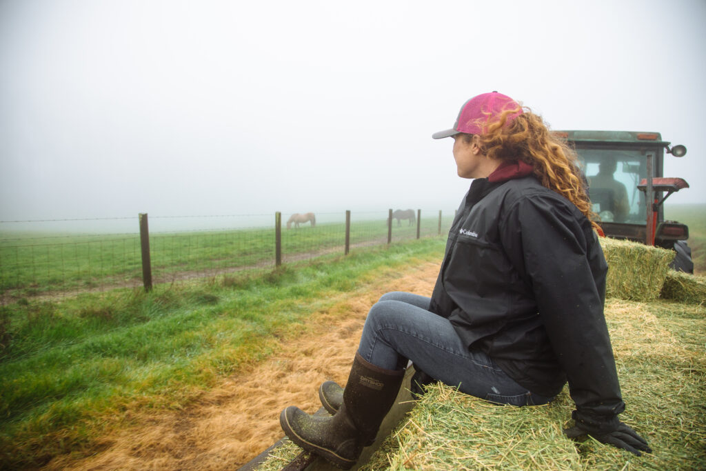 Rancher sitting on hay bales on a tractor-pulled trailer looking out over foggy green pastures with horses visible in the distance at Marshall Home Ranch