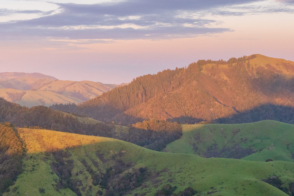 Wide view of rolling green ranchland hills and forested ridgelines at sunset from Spring Valley Ranch, with Hicks Mountain glowing in warm evening light across the protected landscape of Marin County.